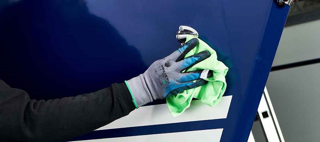 A hand wearing a light gray work glove wipes the side of a dark blue boat with a light green non-woven cloth. The boat is parked on land.