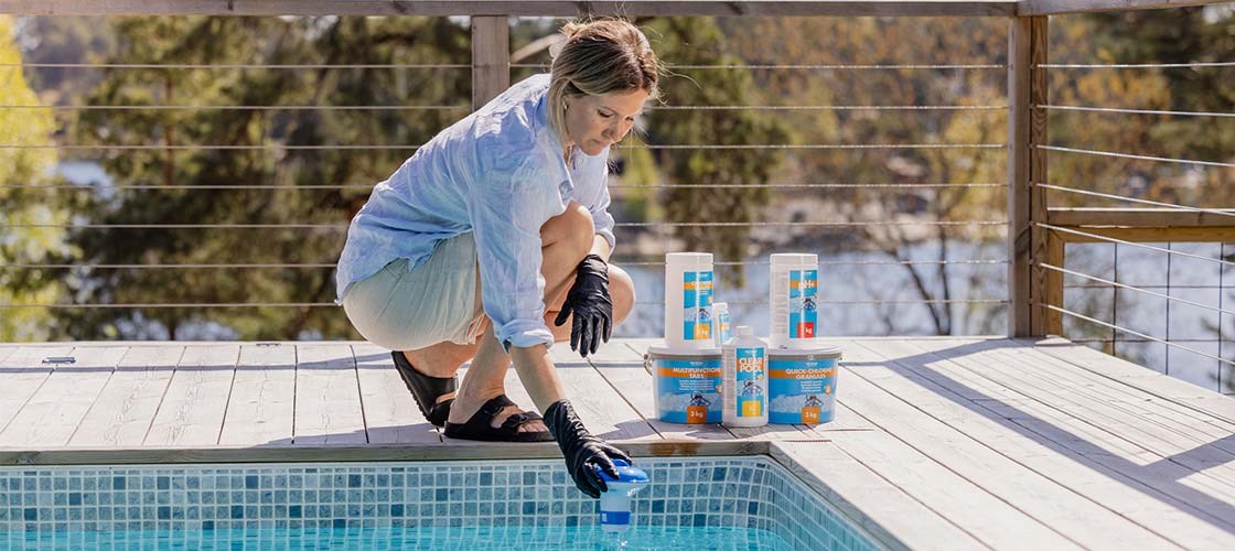 En person hukar vid kanten av en pool på en träterrass och placerar en blå doseringsenhet i vattnet, med flera burkar poolkemikalier uppställda bredvid.