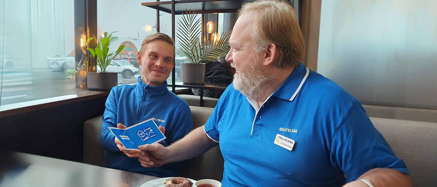 Två personer sitter vid ett bord inomhus. En äldre man med ljust skägg, klädd i en blå pikétröja, tittar leende mot en yngre person i blå tröja bredvid sig. Den yngre personen håller i ett presentkort.