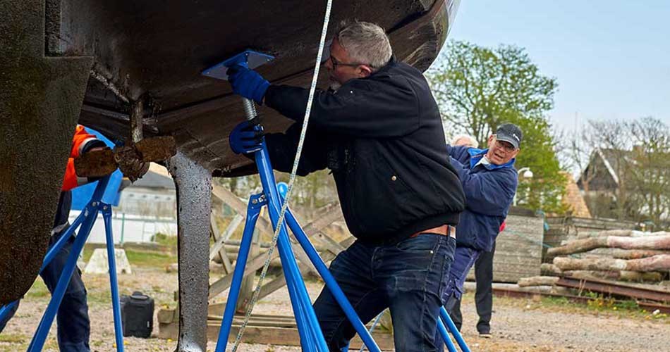 Man arbetar under en båt som är upphöjd med blå båtstöd medan en annan person hjälper till i bakgrunden vid en utomhusbåtbrygga.