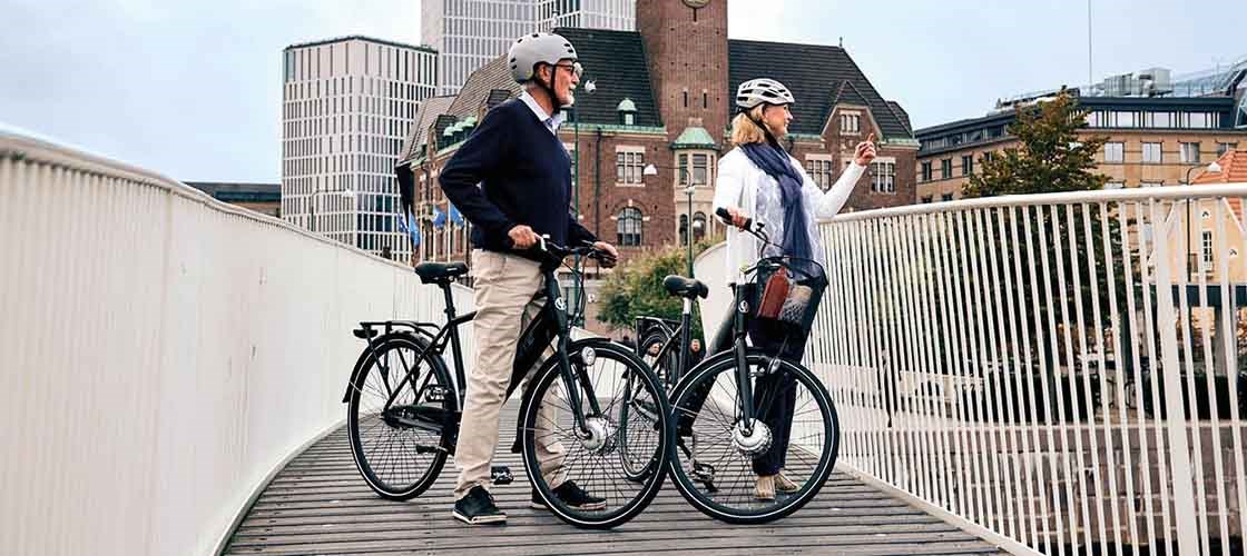 Två cyklister med hjälm och varsin elcykel står på en bro i stadsmiljö och blickar ut.