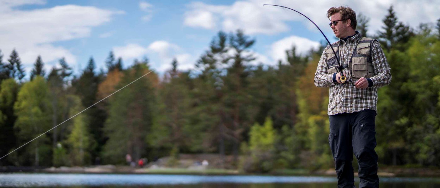En person står vid vattenkanten med ett fiskespö och fiskar i en naturnära miljö. Personen bär rutig skjorta, väst och solglasögon medan fiskespöet är böjt framåt. Träd och grönska syns i bakgrunden under en delvis molnig himmel.