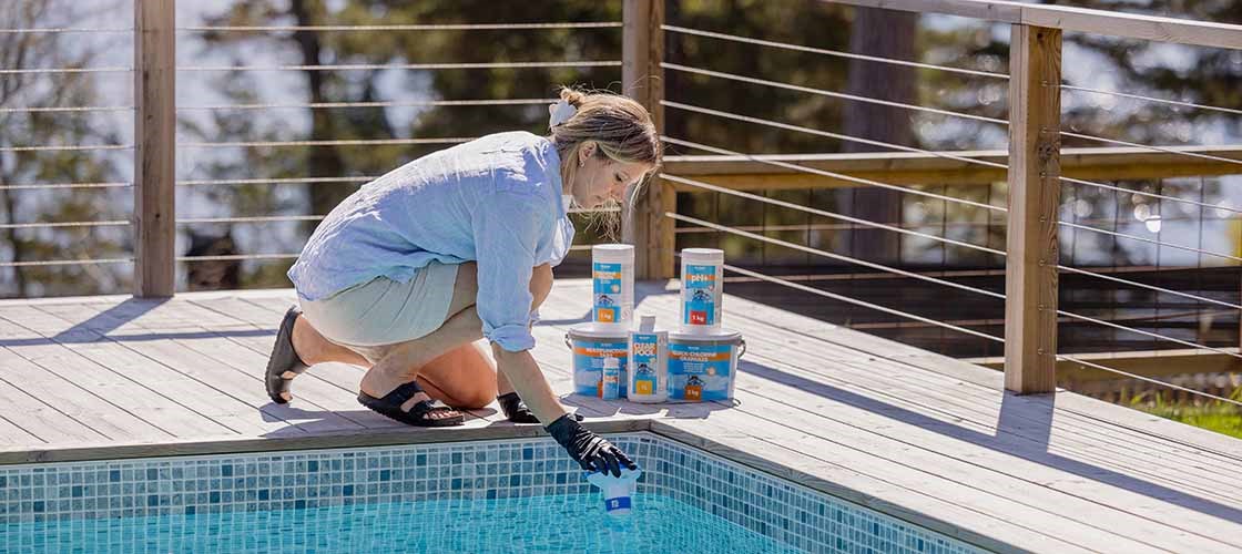 Person i skyddshandske testar poolvattnet vid kanten av en utomhuspool. Flera behållare med poolkemikalier står på trädäcket bredvid poolen.