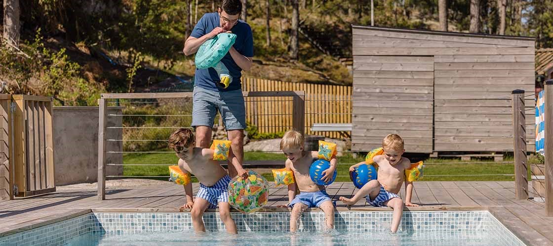 Several people are sitting and standing at the edge of an outdoor swimming pool. Three children are dipping their feet in the pool and an adult man is standing in the background inflating a swimming ring. In the background is a wooden deck, fence, grass area and a small building surrounded by trees. The image shows a summer environment where people are at a pool for swimming and relaxation.