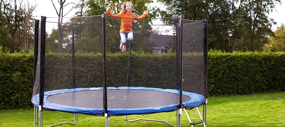 A person jumps on a round trampoline with a safety net that is placed on a lawn in a garden. The trampoline has a ladder on the side and is surrounded by hedges and trees in the background. The image shows an outdoor environment with a focus on the use of a trampoline with a safety net for increased safety.