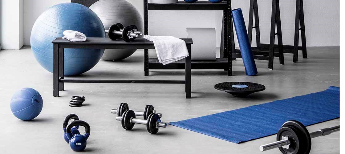 Ett träningsrum med blå och vita träningsredskap, inklusive yogamatta, hantlar, kettlebell, medicinbollar och stora balansbollar. En svart hylla innehåller ytterligare utrustning som vikter, handdukar och träningsband. På väggen syns en handritad tabell med texten ”Squat Challenge”.