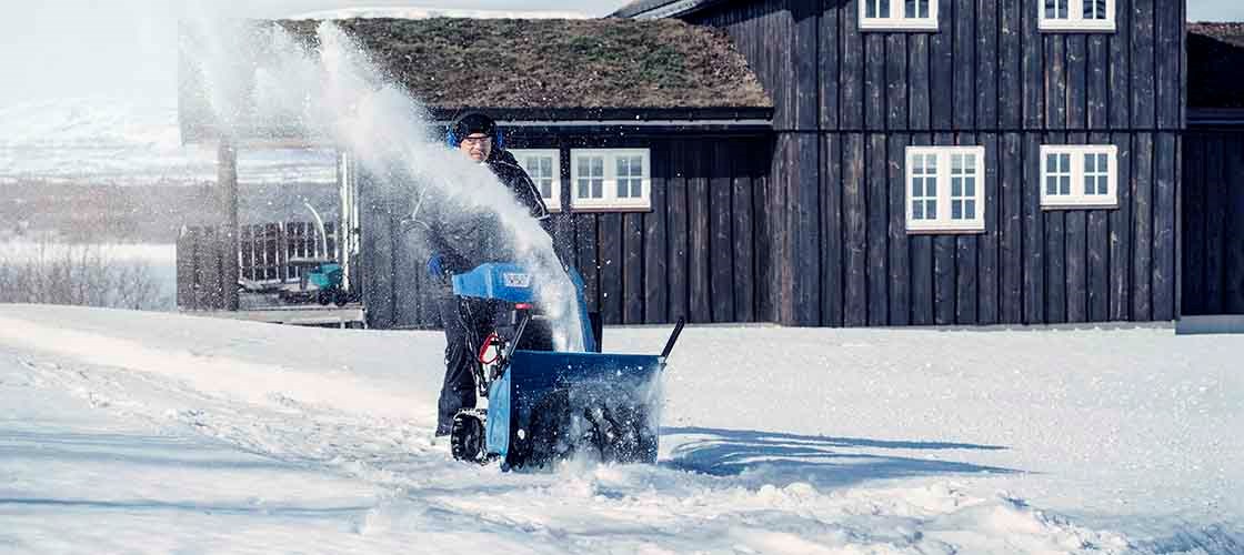 A man dressed in winter clothes, a hat and earmuffs is driving a blue snowblower that is spraying snow. In the background is a dark wooden house and there is a lot of snow on the ground.
