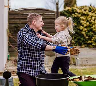 A man wearing gardening gloves hugs a little girl in a garden. In front of them is a pot of soil and in the background is a fence and lush bushes.