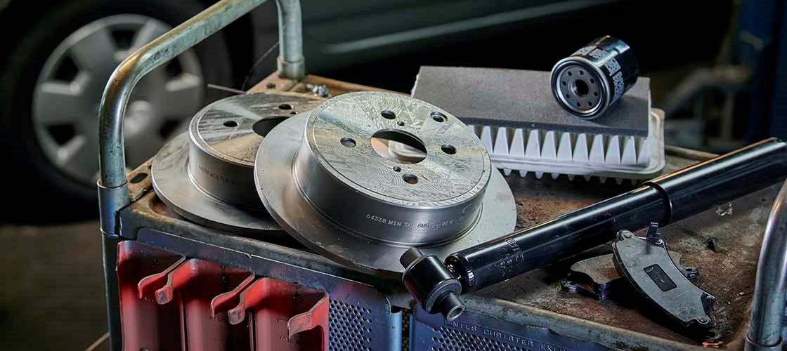 Close-up of a workshop cart with car parts and tools. Two brake discs are stacked next to an oil filter, a drive shaft and a digital measuring tool. In the background, parts of a garage with a car are out of focus.
