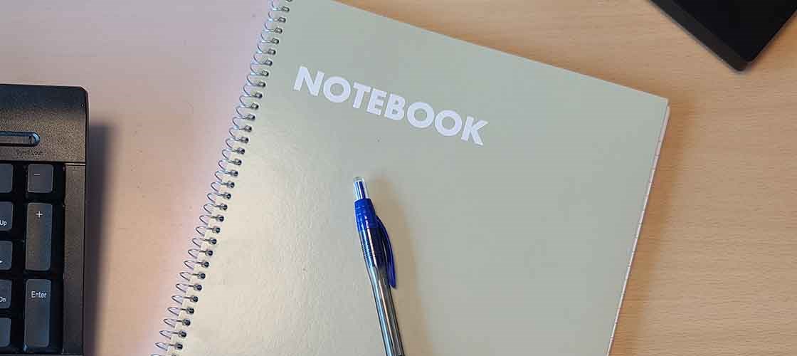 Spiral notebook with the text “NOTEBOOK” and blue ballpoint pen on desk.