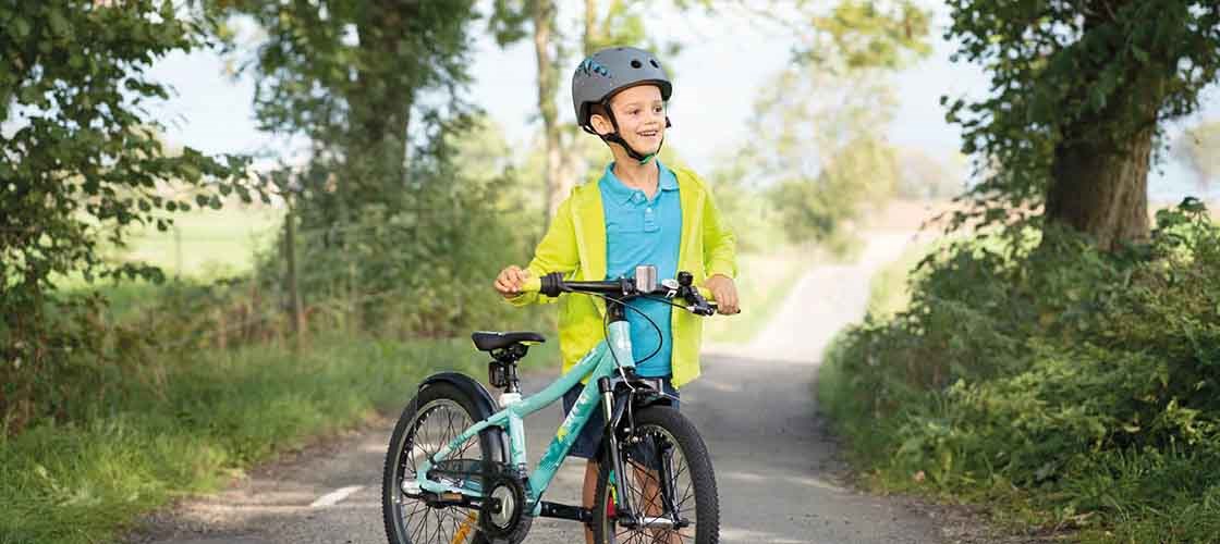 Ett leende barn med cykelhjälm och färgglada kläder står med sin cykel på en solig landsväg omgiven av grönskande träd.
