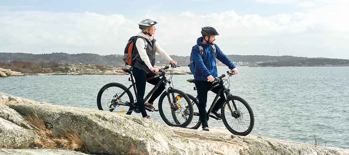Två personer med hjälm står på en klippa vid havet med varsin mountainbike och blickar ut över vattnet.