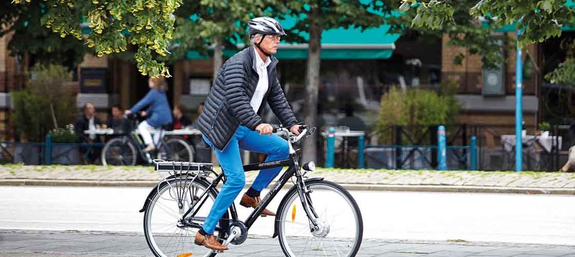 Man with helmet cycling on a city street with shops and trees in the background.
